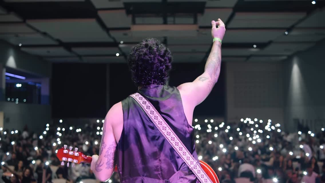 Bruno de costas no palco, de braço erguido, diante da plateia com luzes acesas