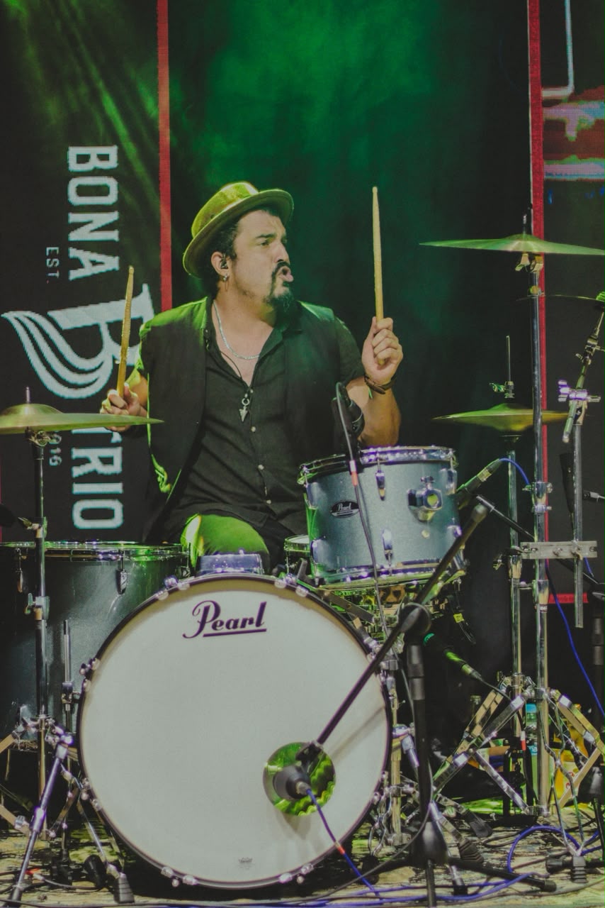 Max na bateria de baquetas erguidas, com banner Bona Trio ao fundo